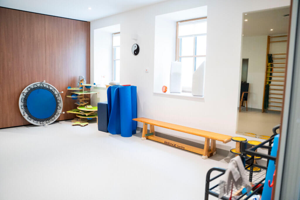 Therapieraum im Gesundheitszentrum mit Trampolin, Matten und Bänken für Physiotherapie.